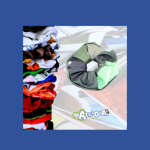 Chouchou d' Atchoum en tissu multicolore à panneaux verts et gris, mise en valeur devant une pile de chouchou de différentes couleurs, avec le logo Atchoum — Artiste rock jeunesse en bas de l'image. Élastique à cheveux
